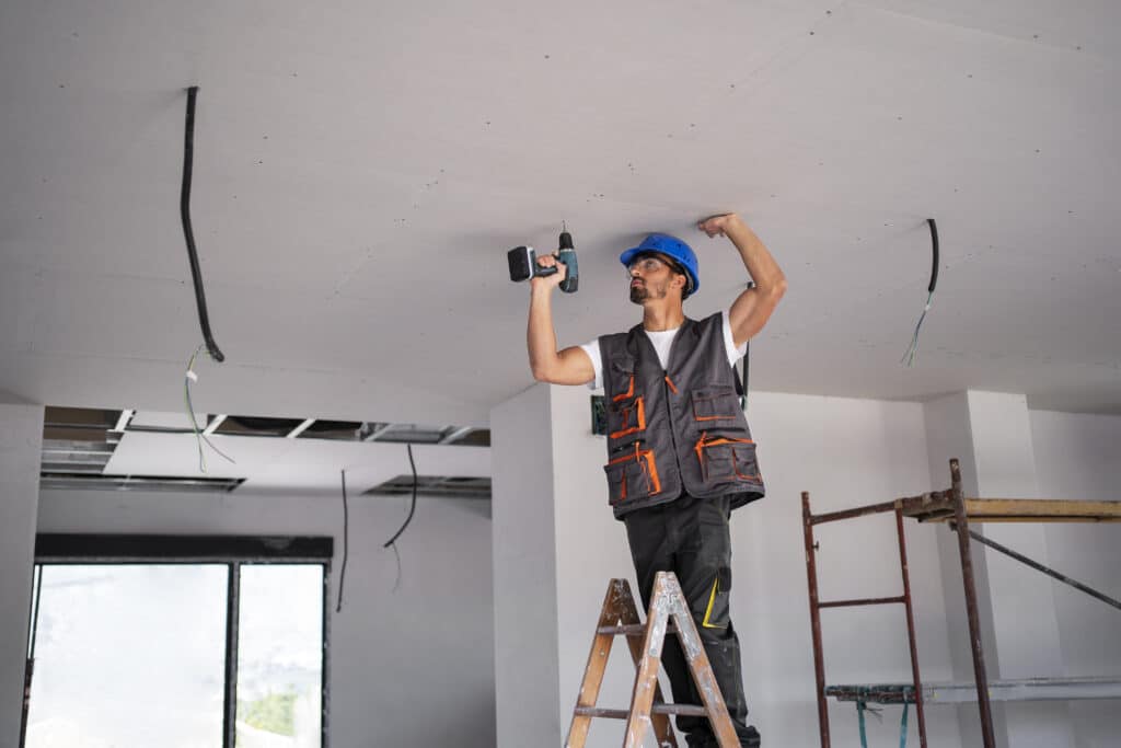 Construction worker installing calcium silicate false ceilings using a power drill, standing on a ladder at an indoor job site with exposed wiring.