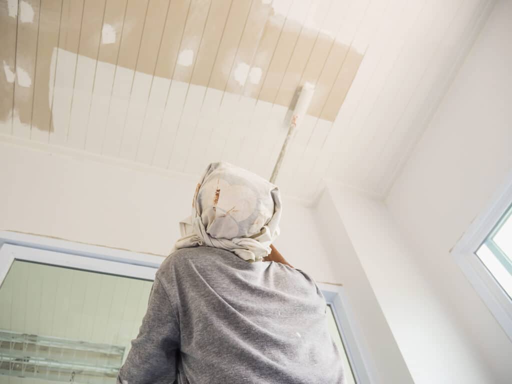 Worker painting calcium silicate false ceiling with a roller brush inside a bright modern room.