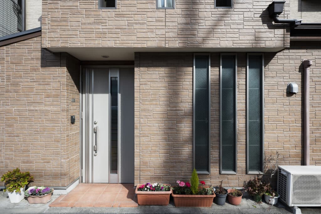 Modern house entrance with vertical windows, metal front door, potted plants, and air conditioning unit on stone exterior wall.