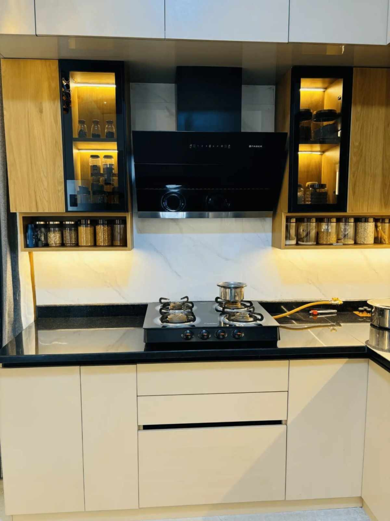 Modern modular kitchen with black granite countertop, built-in gas stove, chimney, wooden cabinets, and illuminated glass storage units.